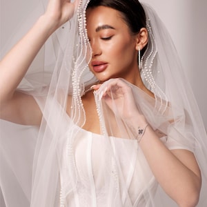 May include: A bride wearing a white wedding dress and veil. The veil is adorned with delicate pearl detailing. The dress features a corset-style bodice and sheer sleeves. The text "PrincessVeil" is visible in the upper right corner.