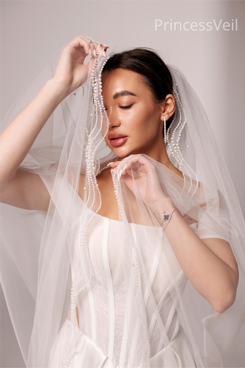May include: A bride wearing a white wedding dress and veil. The veil is adorned with delicate pearl detailing. The dress features a corset-style bodice and sheer sleeves. The text "PrincessVeil" is visible in the upper right corner.