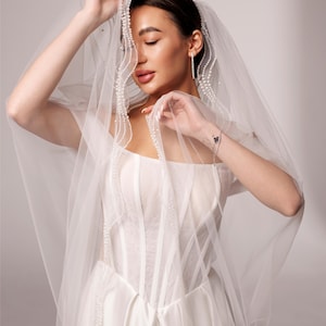 May include: A bride in a white wedding dress with a sheer veil. The veil is adorned with pearls and drapes over the bride's face and shoulders. The dress has a fitted bodice and a flowing skirt. The text "PrincessVeil" is visible in the upper right corner.