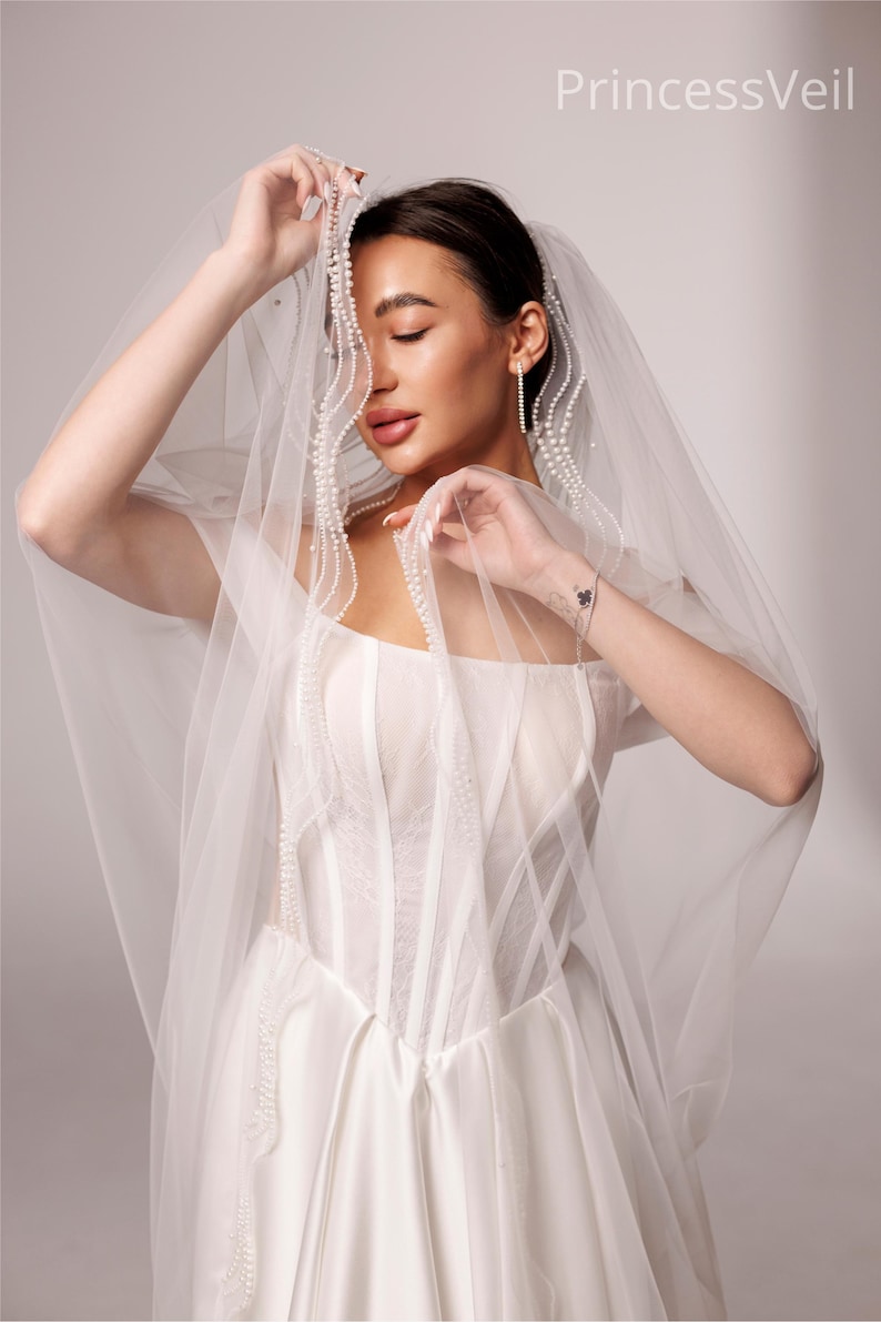 May include: A bride in a white wedding dress with a sheer veil. The veil is adorned with pearls and drapes over the bride's face and shoulders. The dress has a fitted bodice and a flowing skirt. The text "PrincessVeil" is visible in the upper right corner.