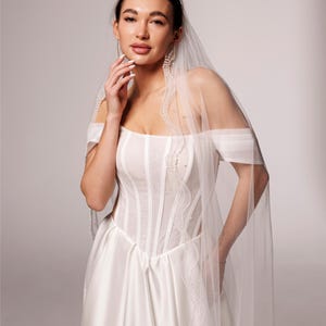 May include: A bride wearing a white wedding dress with a sheer veil. The dress features a fitted bodice with vertical seams and a flowing skirt. The veil is adorned with delicate beading along the edges. The text "PrincessVeil" is visible in the upper right corner.