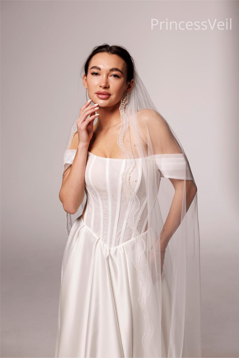 May include: A bride wearing a white wedding dress with a sheer veil. The dress features a fitted bodice with vertical seams and a flowing skirt. The veil is adorned with delicate beading along the edges. The text "PrincessVeil" is visible in the upper right corner.
