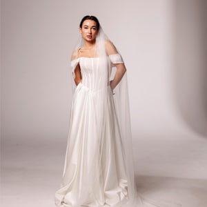 May include: A white satin wedding gown with a fitted bodice and a flowing skirt. The off-the-shoulder design is complemented by a long, sheer veil with a scalloped edge. The text "PrincessVeil" is visible in the upper right corner.