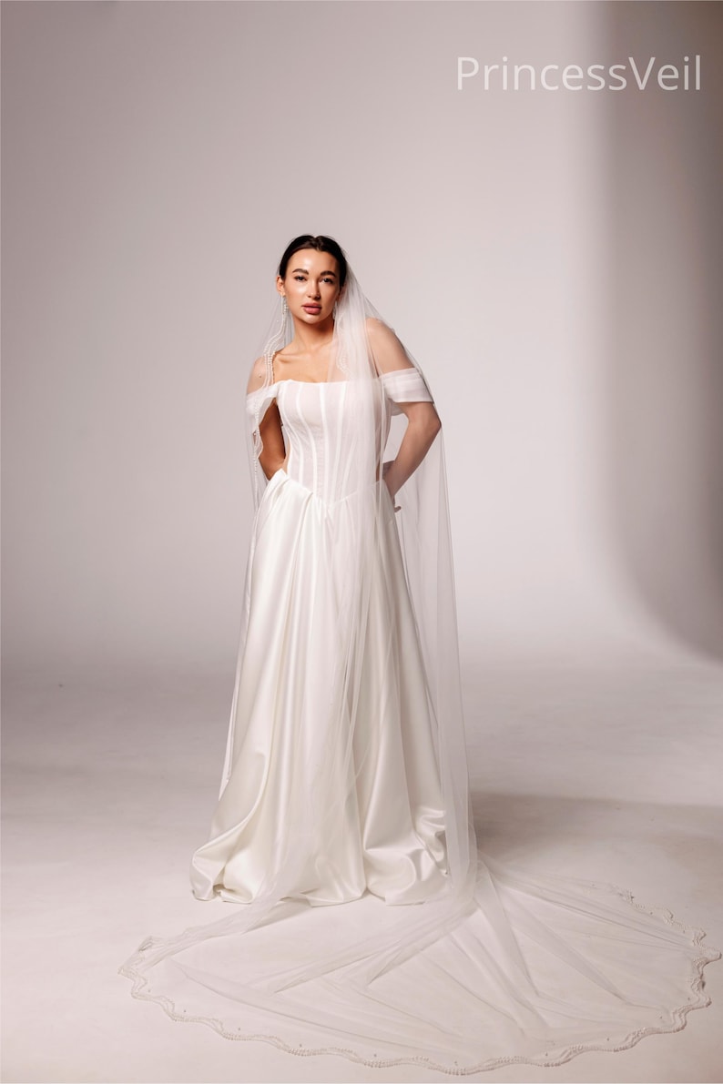 May include: A white satin wedding gown with a fitted bodice and a flowing skirt. The off-the-shoulder design is complemented by a long, sheer veil with a scalloped edge. The text "PrincessVeil" is visible in the upper right corner.