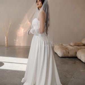 May include: A white wedding dress with a strapless bodice and a flowing skirt. The bride is wearing a white veil with lace trim and a white glove on her left hand.