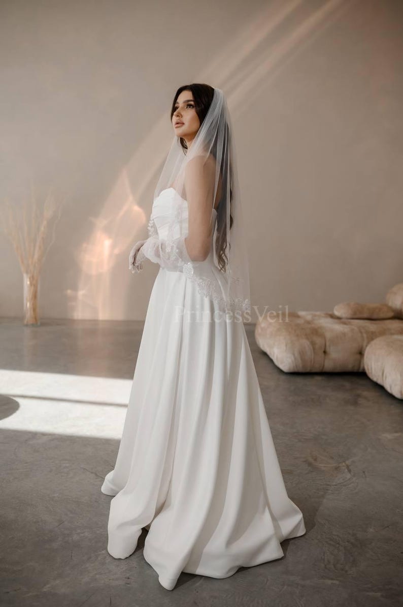 May include: A white wedding dress with a strapless bodice and a flowing skirt. The bride is wearing a white veil with lace trim and a white glove on her left hand.