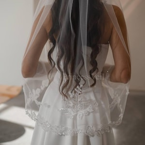 May include: A white bridal veil with a delicate lace trim and beaded embellishments. The veil is worn over a white dress.