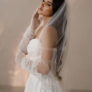 May include: A bride wearing a white wedding dress with a white veil and lace trim. The veil is sheer and has a scalloped edge. The bride is wearing white gloves with glitter.