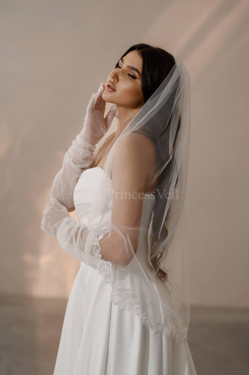 May include: A bride wearing a white wedding dress with a white veil and lace trim. The veil is sheer and has a scalloped edge. The bride is wearing white gloves with glitter.