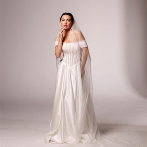 May include: A white satin wedding dress with a corset bodice and off-the-shoulder sleeves. A long, sheer veil with a beaded edge trails behind the dress. The text "PrincessVeil" is in the upper right corner.