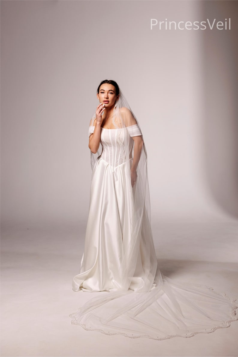 May include: A white satin wedding dress with a corset bodice and off-the-shoulder sleeves. A long, sheer veil with a beaded edge trails behind the dress. The text "PrincessVeil" is in the upper right corner.