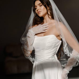 May include: A bride wearing a white strapless wedding dress with a veil and long white gloves with sparkly embellishments.