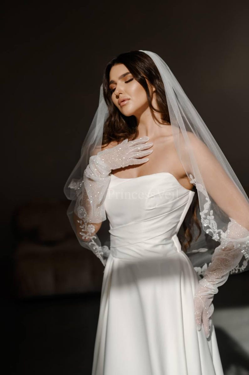 May include: A bride wearing a white strapless wedding dress with a veil and long white gloves with sparkly embellishments.