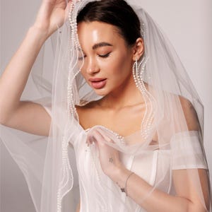 May include: A sheer white bridal veil with a delicate pearl trim, held up by a person. The veil is draped over a white wedding dress. The text "PrincessVeil" is visible in the upper right corner.