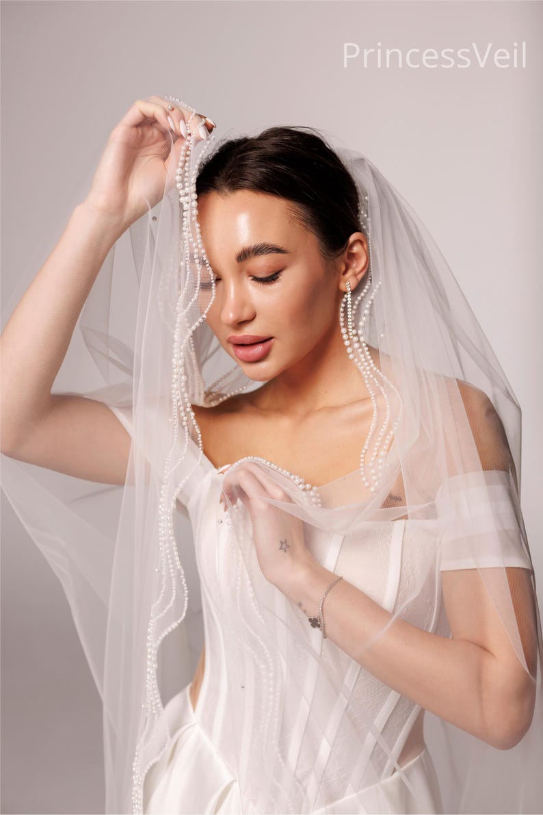May include: A sheer white bridal veil with a delicate pearl trim, held up by a person. The veil is draped over a white wedding dress. The text "PrincessVeil" is visible in the upper right corner.