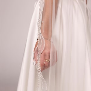 May include: A sheer white wedding veil with a delicate pearl trim. The veil is held up, revealing the intricate pearl detailing and the text "PrincessVeil". The veil is paired with a white satin wedding dress.