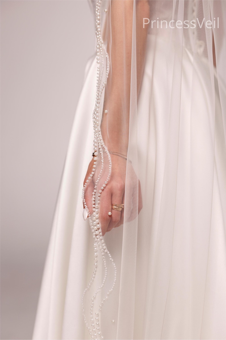 May include: A sheer white wedding veil with a delicate pearl trim. The veil is held up, revealing the intricate pearl detailing and the text "PrincessVeil". The veil is paired with a white satin wedding dress.