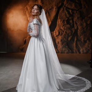 Könnte beinhalten: Ein weißes Brautkleid mit einem langen, fließenden Rock und einem Spitzenoberteil. Das Kleid wird mit einem langen, weißen Schleier mit Spitzenbesatz kombiniert. Die Braut steht in einer dunklen, rustikalen Umgebung.