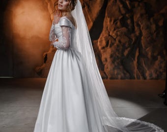 May include: A white wedding dress with a long, flowing skirt and a lace bodice. The dress is paired with a long, white veil with a lace trim. The bride is standing in a dark, rustic setting.