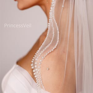 May include: A white bridal veil with a delicate pearl trim. The veil is sheer and drapes softly, with the text "PrincessVeil" visible. The pearls are small and evenly spaced along the edge, creating an elegant look.