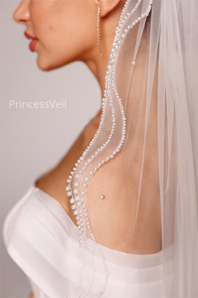 May include: A white bridal veil with a delicate pearl trim. The veil is sheer and drapes softly, with the text "PrincessVeil" visible. The pearls are small and evenly spaced along the edge, creating an elegant look.