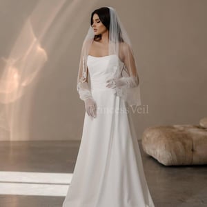 May include: A white wedding dress with a strapless bodice and a flowing skirt. The bride is wearing a white veil with lace trim and white gloves. The dress is photographed against a light brown background.