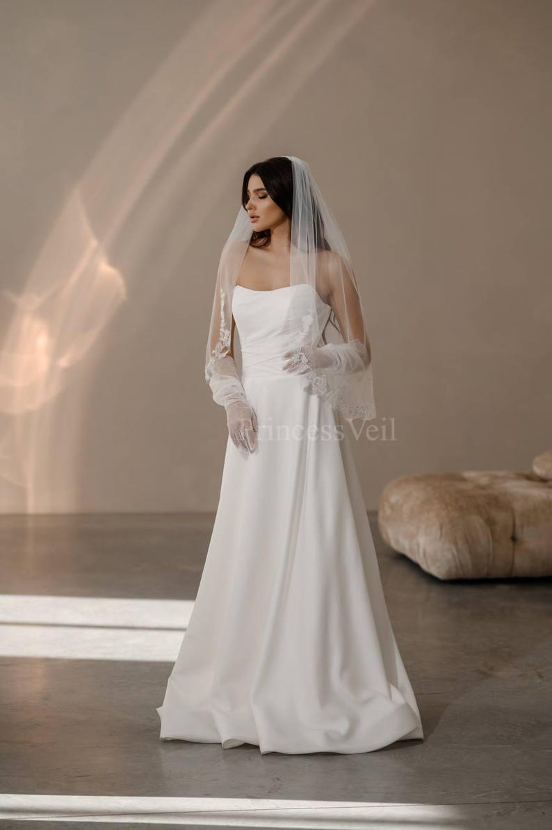 May include: A white wedding dress with a strapless bodice and a flowing skirt. The bride is wearing a white veil with lace trim and white gloves. The dress is photographed against a light brown background.