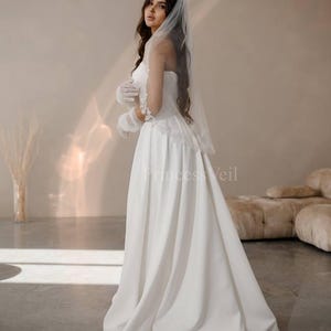 May include: A white wedding dress with a lace bodice and a flowing skirt. The dress is paired with a white veil and gloves.