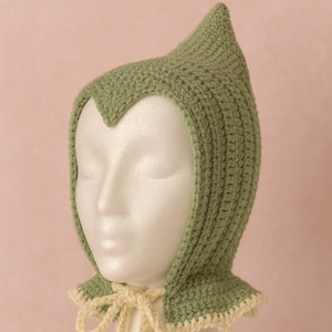 Puede incluir: Un gorro de ganchillo verde salvia con una corona puntiaguda y un ribete festoneado color crema. El gorro está atado con hilo color crema. El gorro se exhibe en una cabeza de maniquí blanca.