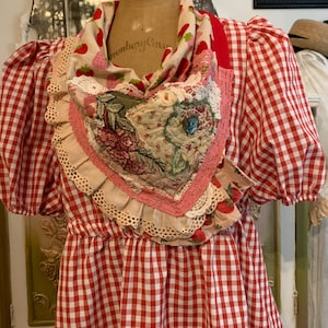 Puede incluir: Vestido de cuadros rojos y blancos con un babero en forma de corazón. El babero presenta tela floral y de fresas, ribete de encaje y un botón azul. El vestido tiene mangas cortas y abullonadas y una cintura ajustada.