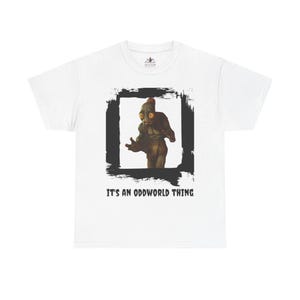 May include: A white short-sleeved t-shirt featuring a central graphic of a brown, large-eyed creature inside a black brushstroke frame. Below the graphic, the text reads IT'S AN ODDWORLD THING in a bold, black, distressed-style sans-serif font.