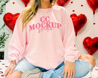 May include: A light pink sweatshirt with the text "CC Mockup Studio" in pink. The model is wearing light blue jeans and gray boots. Red heart-shaped balloons are in the background.
