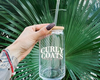 Op de afbeelding: Een heldere glazen beker met een bamboe deksel en een helder rietje. De beker heeft de tekst "CURLY COATS mockup studio" in het wit. De achtergrond is een groen palmblad.