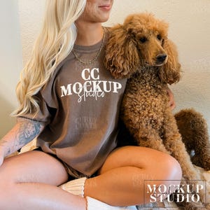 Puede incluir: Una mujer lleva una camiseta marr&oacute;n con el texto "CC MOCKUP Studio" en blanco. Est&aacute; sosteniendo un caniche marr&oacute;n.