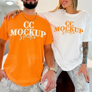 May include: Two people wearing matching t-shirts. One shirt is orange, the other is white. Both shirts have the text "CC Mockup Studio" in a bold font. The models are wearing patterned pants.