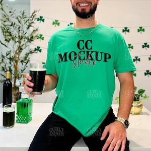 May include: A green t-shirt with the text "CC MOCKUP Studios" in black. The person is holding a glass of dark beer. The background includes shamrock decorations and a bottle of wine.