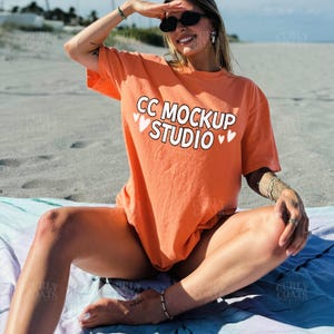 May include: An orange t-shirt with the text "CC MOCKUP STUDIO" in white, accented with small white hearts. The shirt is worn on a beach. The model is wearing sunglasses and jewelry.