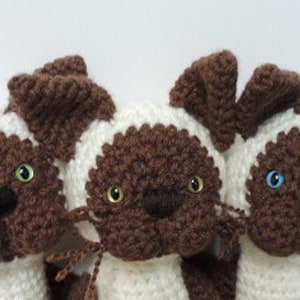 May include: Three crocheted Siamese cat plushies with brown and white fur. The cats have different colored eyes, one with green, one with blue, and one with both green and blue.