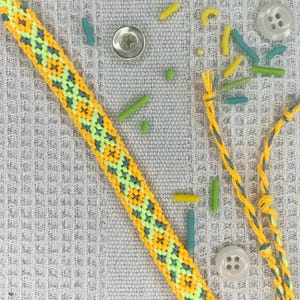 May include: Handmade friendship bracelet in yellow, green, and blue geometric pattern. The bracelet is made of woven thread and has a braided closure. The background includes decorative elements like buttons and sprinkles.