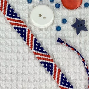 May include: A handmade friendship bracelet in red, white, and blue, with a woven pattern. The image also features red and white buttons, blue beads, and star-shaped embellishments, all arranged on a white textured surface.