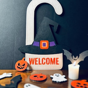 May include: Wooden Halloween door hanger featuring a black witch hat with a purple band and orange buckle, and the word "WELCOME" in orange. Includes pumpkin and bat cutouts, and a lit candle. Festive decor.