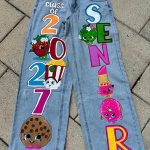 Può includere: Jeans in denim azzurro con disegni dipinti a mano. La gamba sinistra mostra la scritta Class of 2027 con illustrazioni di una fragola, popcorn e un biscotto. La gamba destra presenta la parola SENIOR con una mela verde, un rossetto e una ciambella.