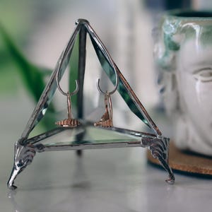 May include: A silver triangular display stand with two gold earrings on it. The stand has a glass top and ornate metal feet.
