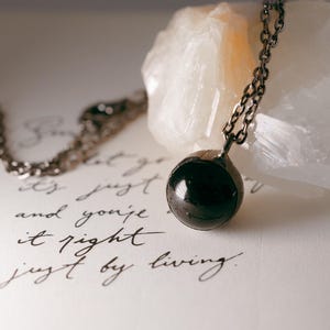 May include: A silver chain necklace with a black gemstone pendant. The pendant is round and has a smooth, polished surface. The necklace is lying on a white surface with a handwritten message.