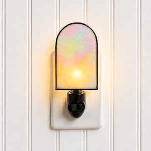 May include: An arched night light featuring iridescent stained glass with a soft, warm glow. The glass panel is framed in black metal and mounted on a black plug-in base with a small toggle switch, emitting a gentle light through the translucent surface.