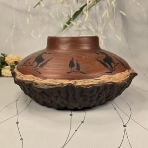 May include: A brown ceramic vase with a textured, dark brown base and a lighter brown upper section. The vase features black silhouette designs of animals. The rim is a light tan color.