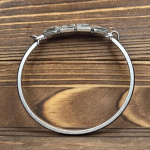 Puede incluir: Pulsera plateada con una pieza central rectangular con gemas. La pulsera tiene un dise&ntilde;o articulado y una banda circular simple. Las gemas son transparentes y est&aacute;n engastadas en una fila, agregando un toque de brillo al accesorio. La pulsera se muestra sobre un fondo de madera.