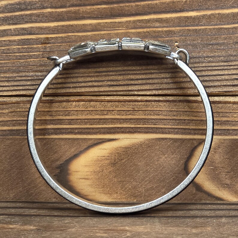 Puede incluir: Pulsera plateada con una pieza central rectangular con gemas. La pulsera tiene un dise&ntilde;o articulado y una banda circular simple. Las gemas son transparentes y est&aacute;n engastadas en una fila, agregando un toque de brillo al accesorio. La pulsera se muestra sobre un fondo de madera.