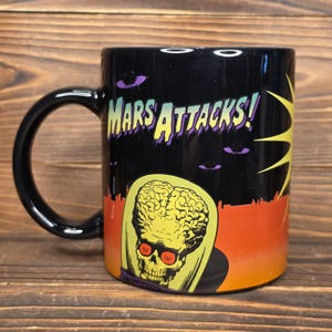 May include: Black ceramic mug featuring the text "MARS ATTACKS!" in yellow and purple. The mug has a graphic of a Martian with a large brain and red eyes. The background includes flying saucers and a cityscape.