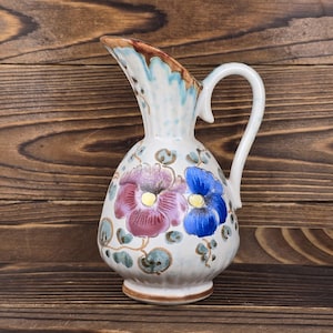 May include: A ceramic pitcher with a white base, adorned with purple and blue floral designs. The pitcher has a curved handle and a spout with brown and blue accents. The background is a wooden surface.